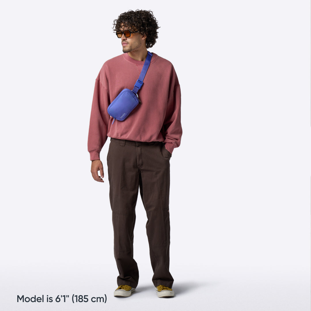 Owala Daily Crossbody in Rest Azured, periwinkle blue belt bag worn crossbody by a man in a red sweatshirt and brown pants, with text reading "Model is 6'1" (185 cm)"