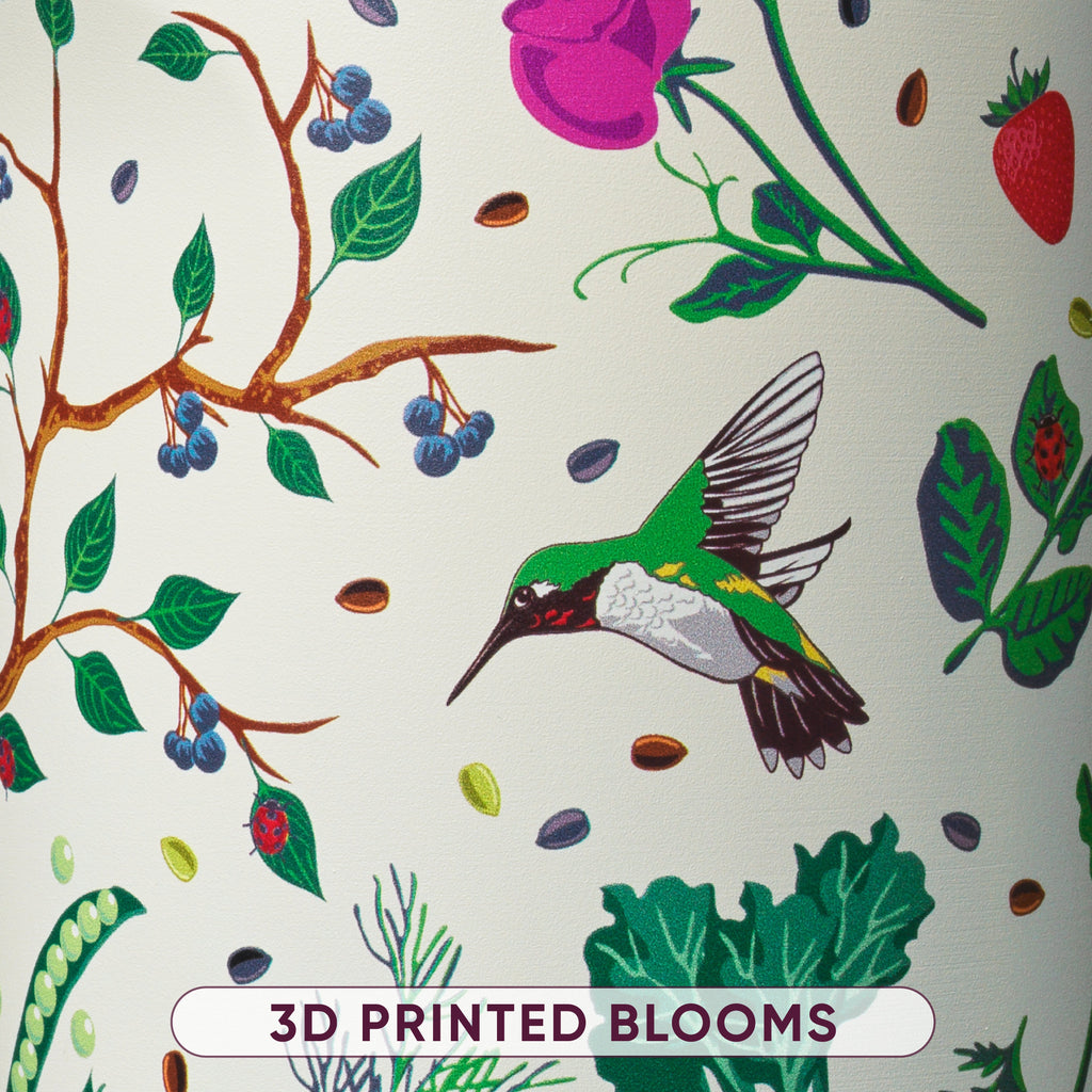 Close-up of cream bottle body printed with hummingbird, blueberry branches, pink rose, ladybug, and leafy plant illustrations, labeled 3D Printed Blooms
