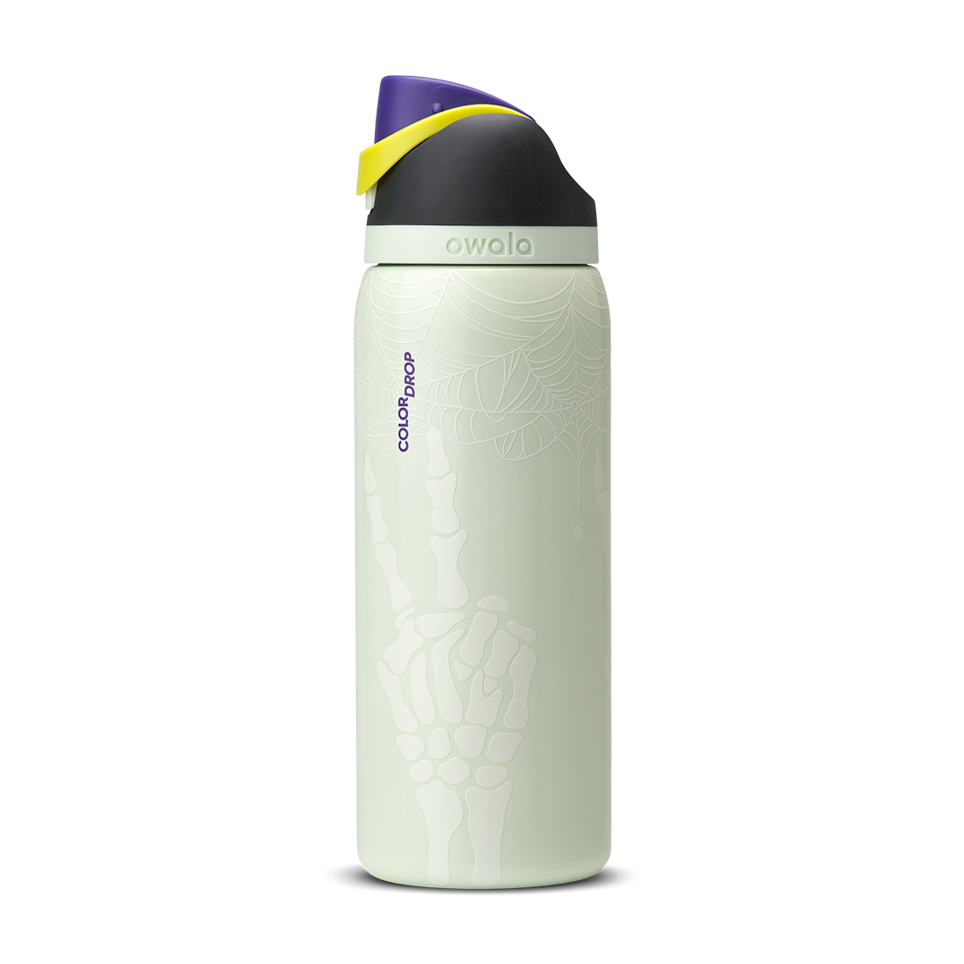 Owala FreeSip 32oz in RIP, pale mint green insulated stainless steel bottle with white skeleton-hand pattern body, dark charcoal lid, yellow carry loop, and purple flip cap, Color Drop branding on body; glows in the dark