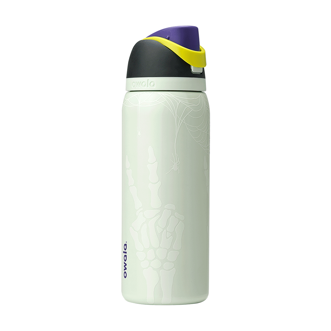 Owala FreeSip 32oz in RIP, flip cap open showing yellow-green FreeSip spout, purple lid interior with gray silicone seal, dark charcoal lid, on pale mint green body with white skeleton-hand pattern; glows in the dark